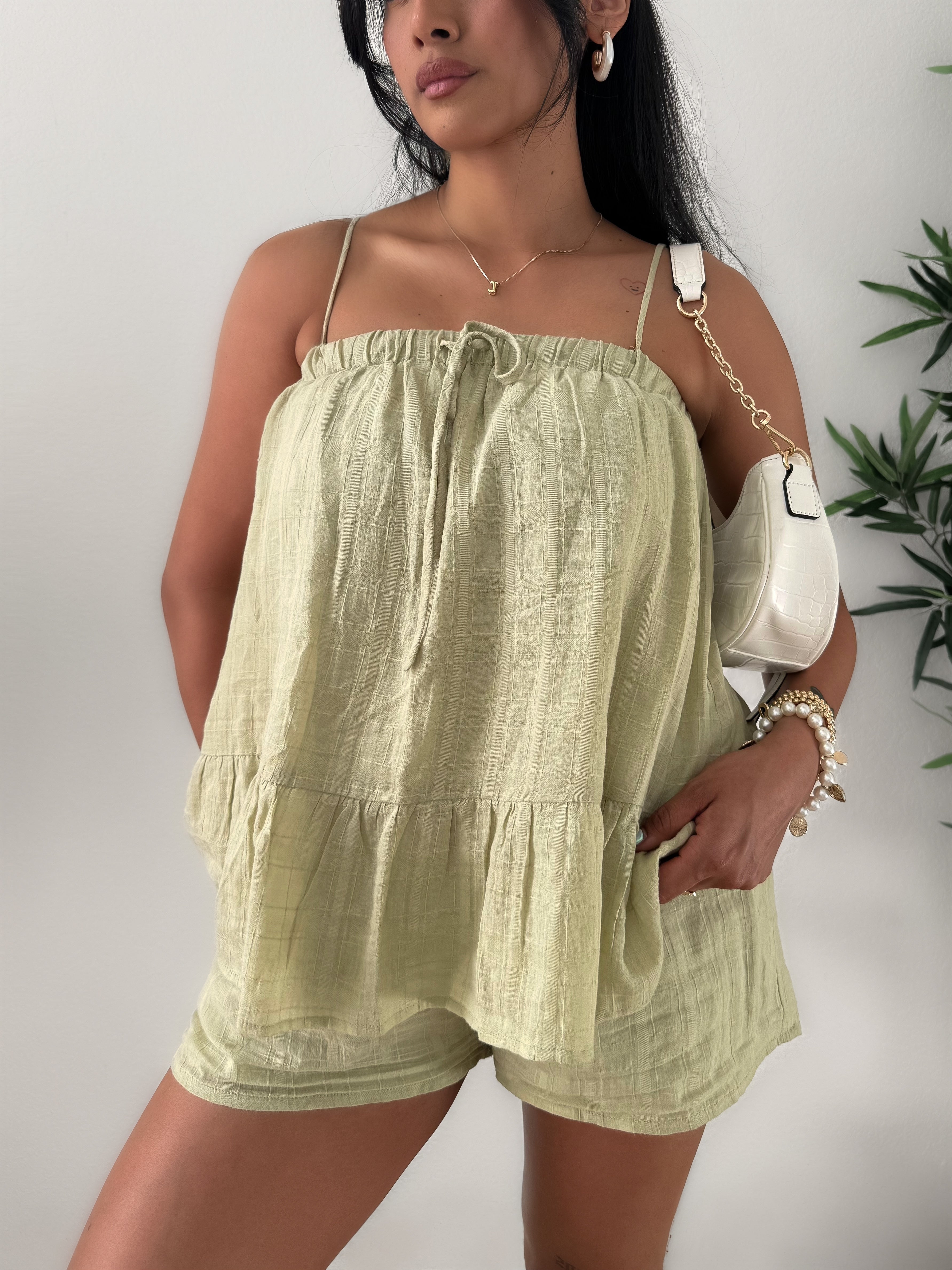 Woman wearing a light green tiered top and shorts with a white handbag against a plain background